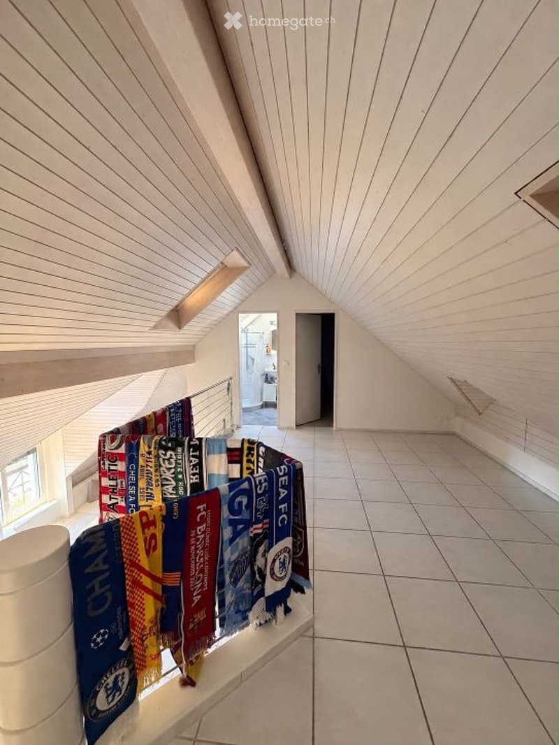 An attic space with exposed wooden beams, white walls, and tiled flooring. A small window and a doorway lead to another room. A collection of scarves is displayed on a stand near the entrance.