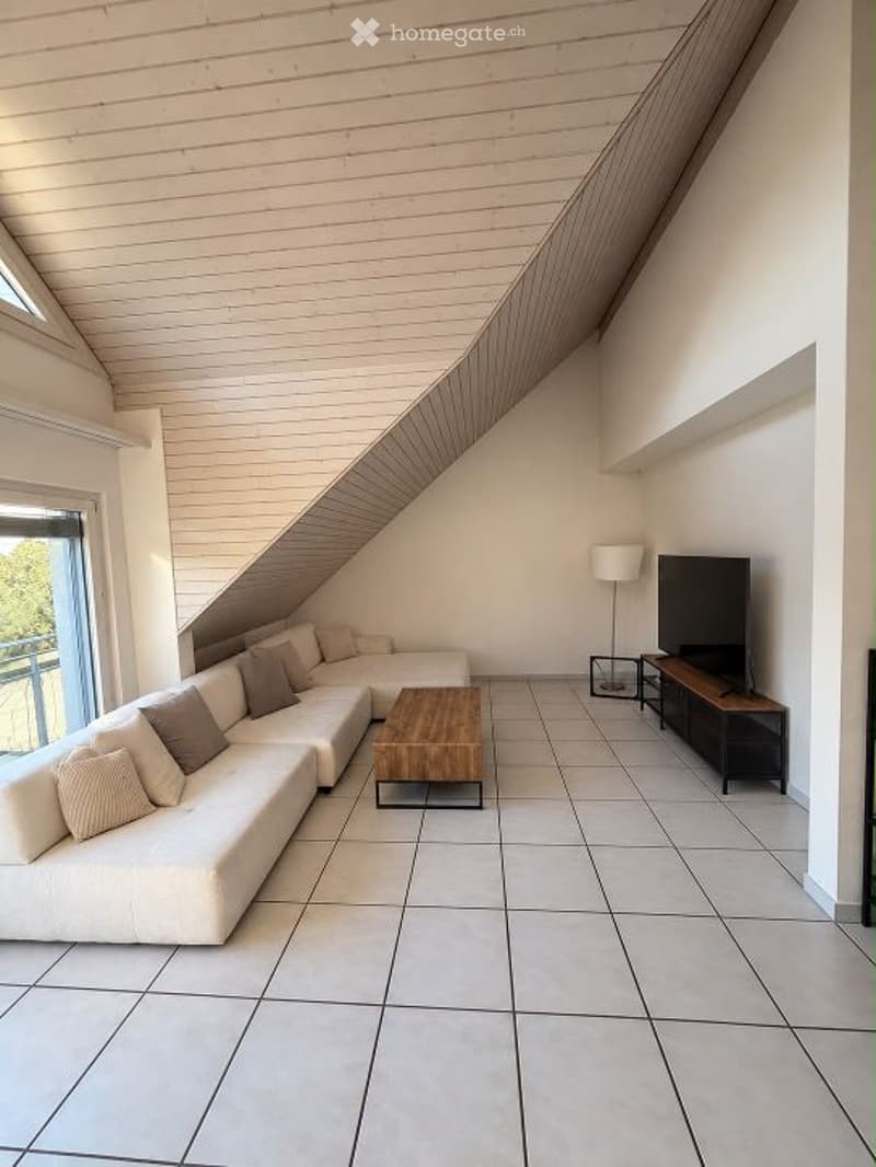 Modern living room with white walls, light wood ceiling beams, large windows, and a balcony. Features a white sectional sofa, wooden coffee table, floor lamp, and TV on a stand.
