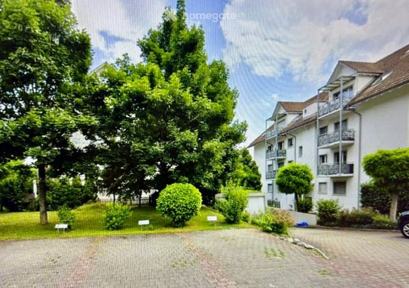 Multi-story residential building with balconies, surrounded by greenery and trees. The building has a white facade and is situated in a well-maintained area with paved pathways.