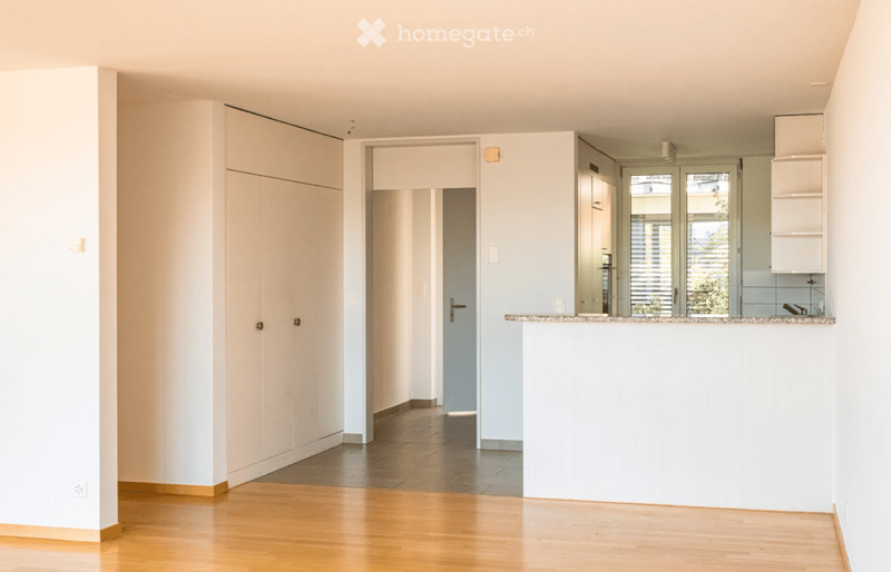 Bright, open-plan living and dining area with wooden flooring, white walls, and built-in storage units. A doorway leads to another room, and a kitchen area is visible with a countertop and cabinets.