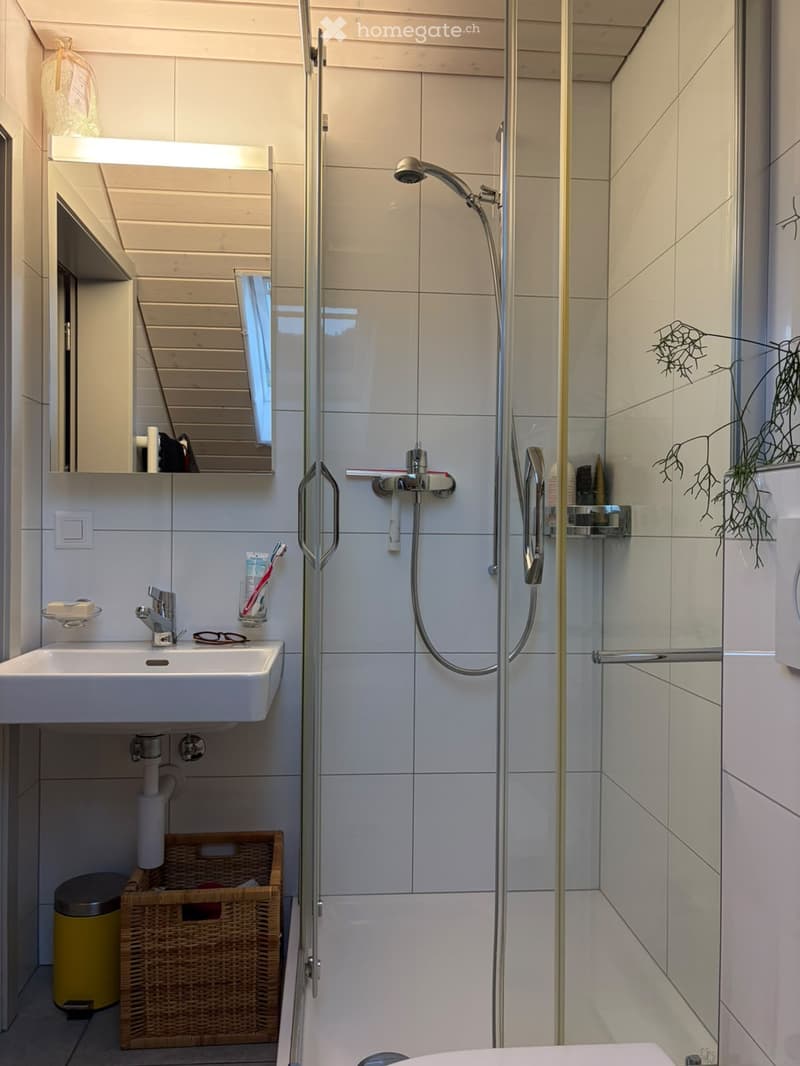 Modern bathroom with white tiles, a sink with a mirror, a shower with a glass enclosure, and a wicker basket on the floor.