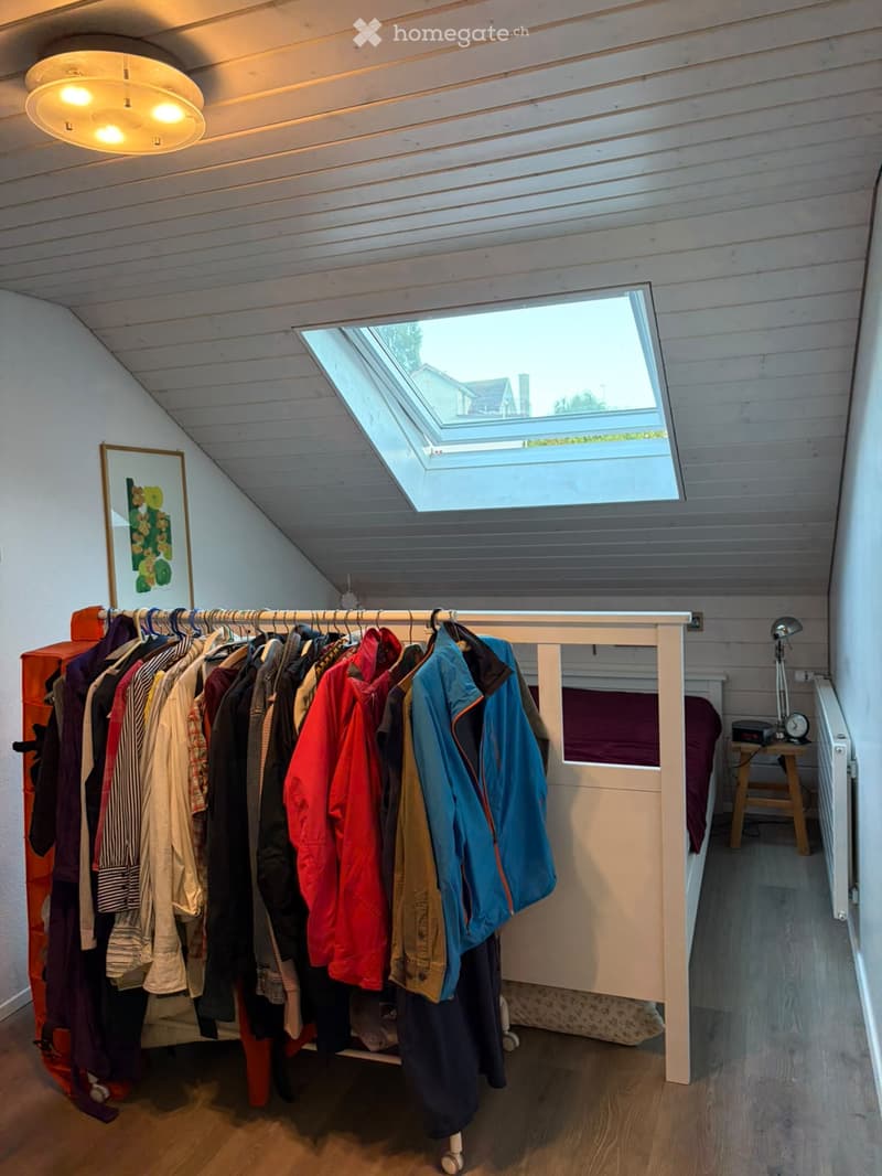 A small, cozy attic room with wooden walls and ceiling, featuring a lofted bed, a clothing rack, a picture frame, and a small table with a lamp.