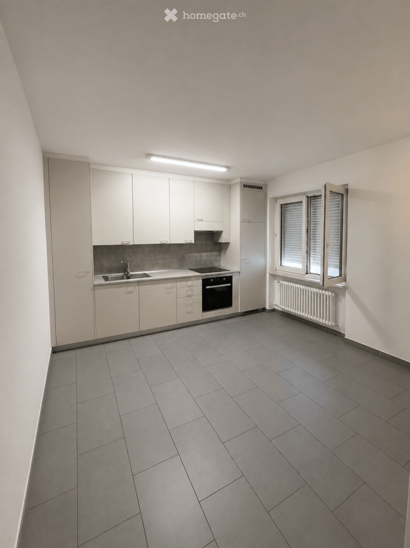 Modern kitchen with grey tiled floor, white cabinets, sink, oven, and radiator under window.