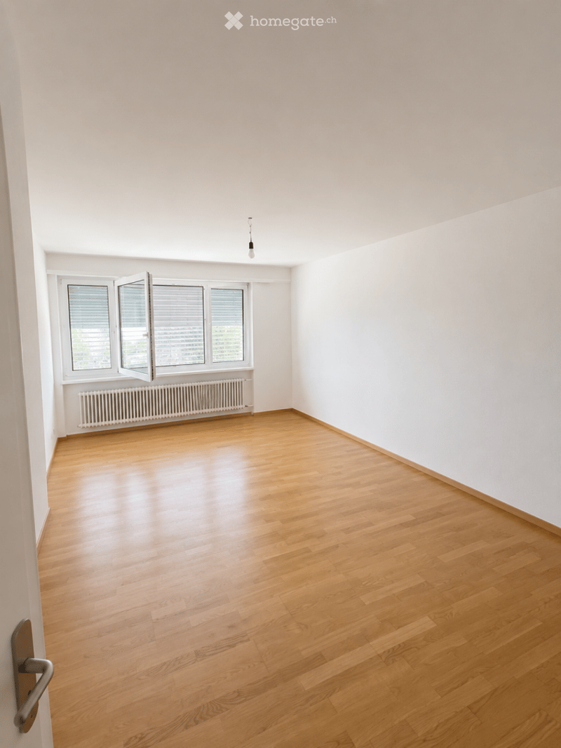 Empty room with wooden flooring, white walls, and large windows with blinds. A radiator is visible below the windows, and a single light fixture hangs from the ceiling.