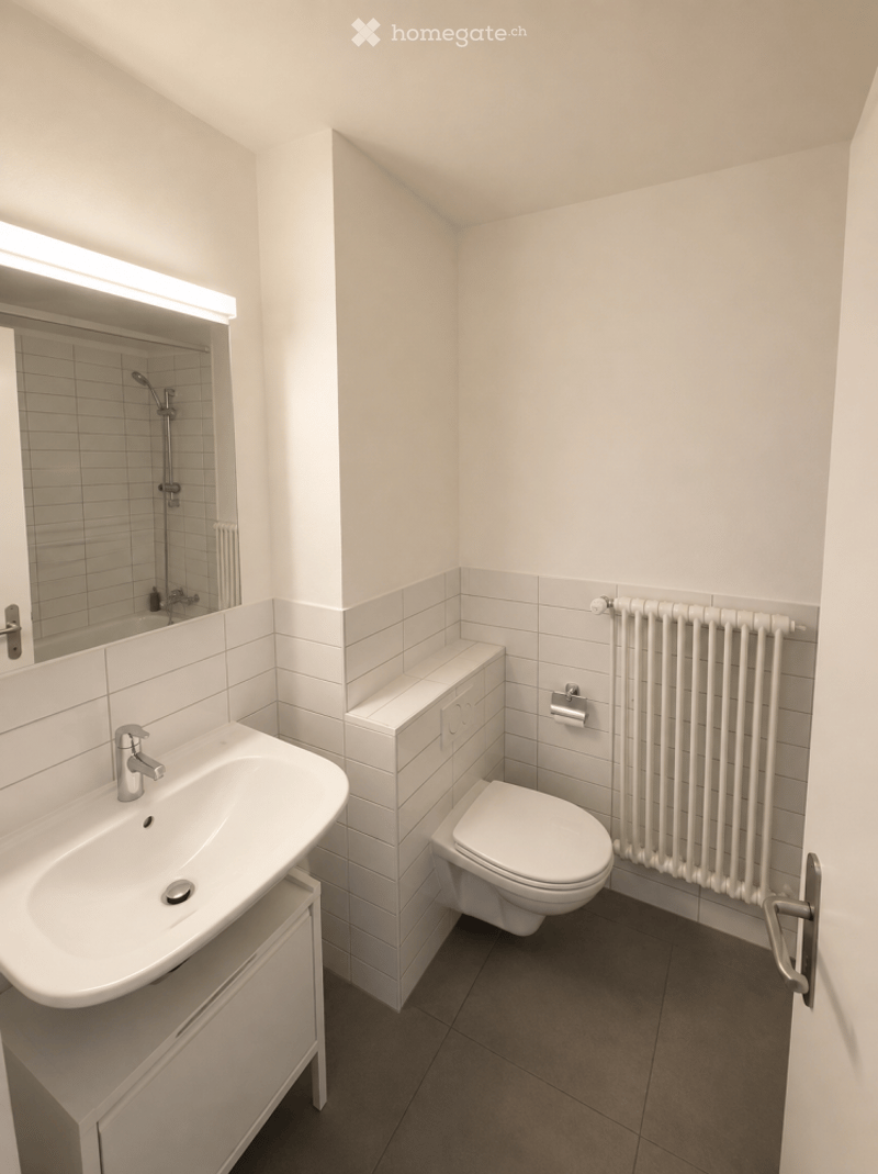 Modern, compact bathroom with white tiles, a sink, toilet, radiator, and a shower area with a mirror above it.