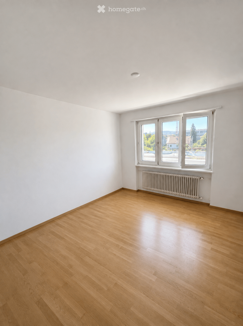 Empty room with light wooden flooring, white walls, and large windows offering a view of the outside. No furniture or equipment is present.