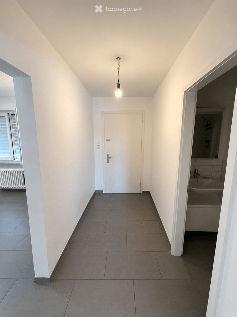 Empty hallway with white walls, tiled floor, and a single light fixture. A door leads to another room, and a window is visible on the left side.