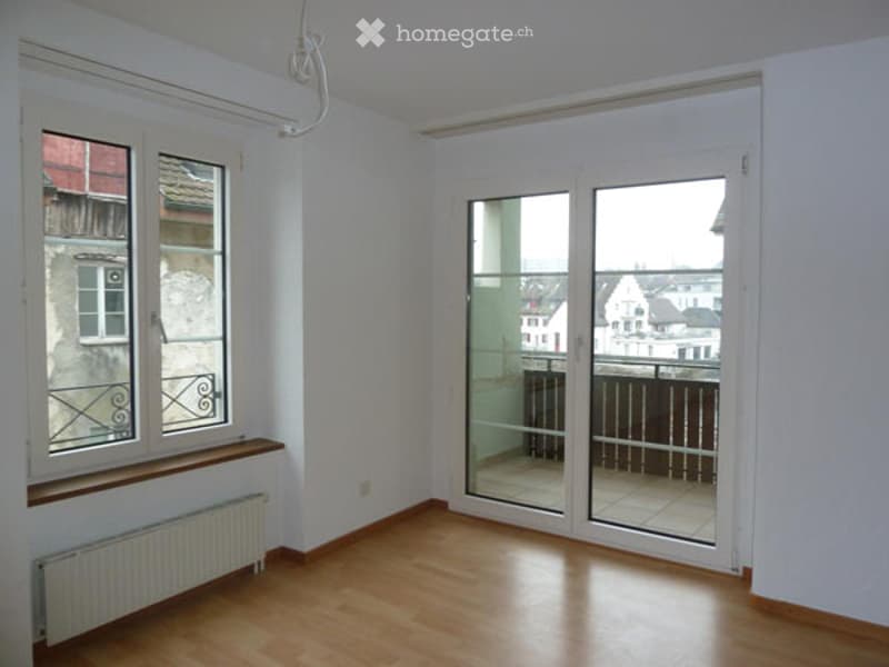 Empty room with wooden flooring, white walls, large window with wooden sill, and glass sliding doors leading to a balcony.