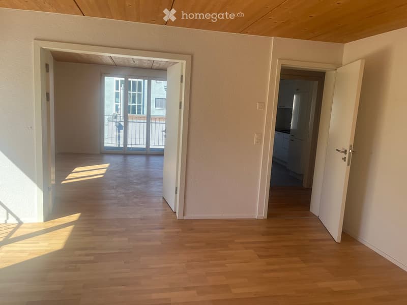 Empty hallway with wooden flooring, white walls, and open doors leading to a balcony and a kitchen area.