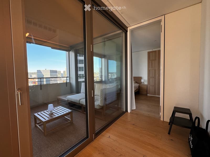 Modern balcony with a view of the city, featuring a small table with a lamp, a stool, and a sofa, connected to a bedroom with wooden flooring and a bed.