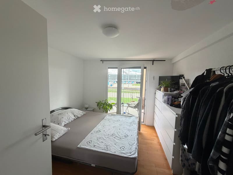 Bedroom with a double bed, nightstand, drawer unit, clothing rack, small table, and chair visible through the open sliding door leading to a balcony.