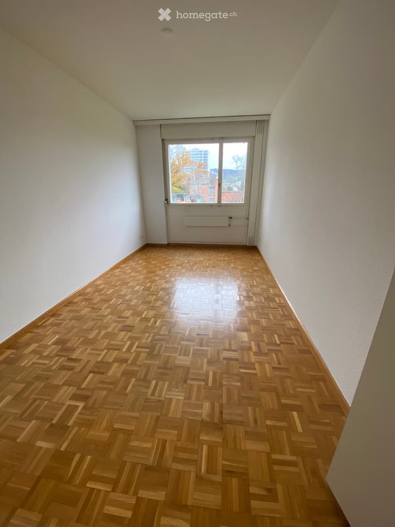 Empty hallway with wooden herringbone flooring, white walls, and a large window at the end showing a view of buildings and trees outside.