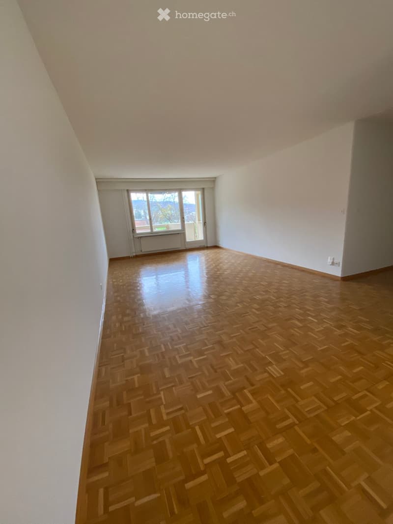 Empty room with wooden parquet flooring, white walls, and a large window with a door leading to a balcony.