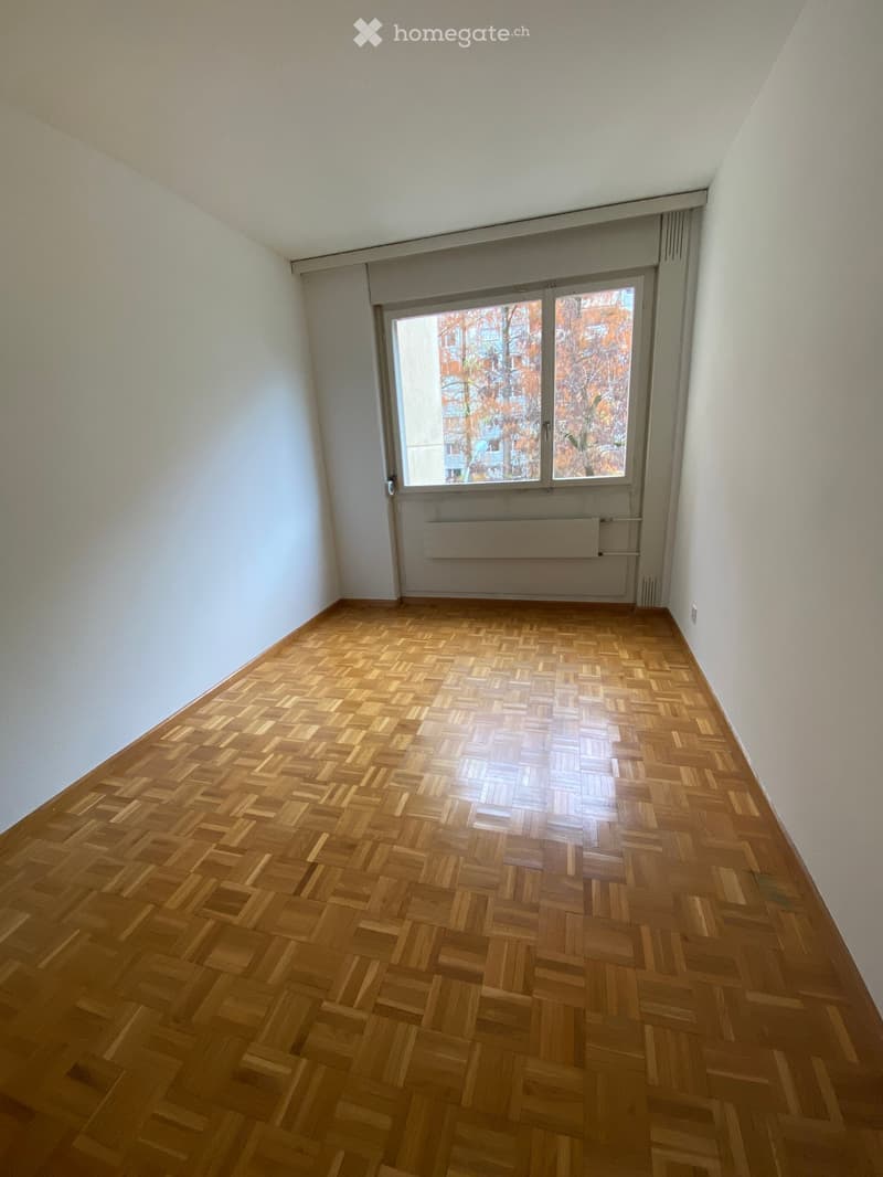 Empty room with wooden herringbone flooring, white walls, and a large window with blinds. No furniture or equipment is present.