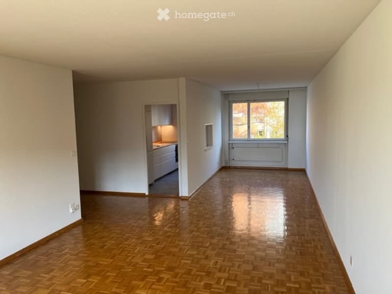 Empty room with wooden parquet flooring, white walls, and a doorway leading to a small kitchen area.