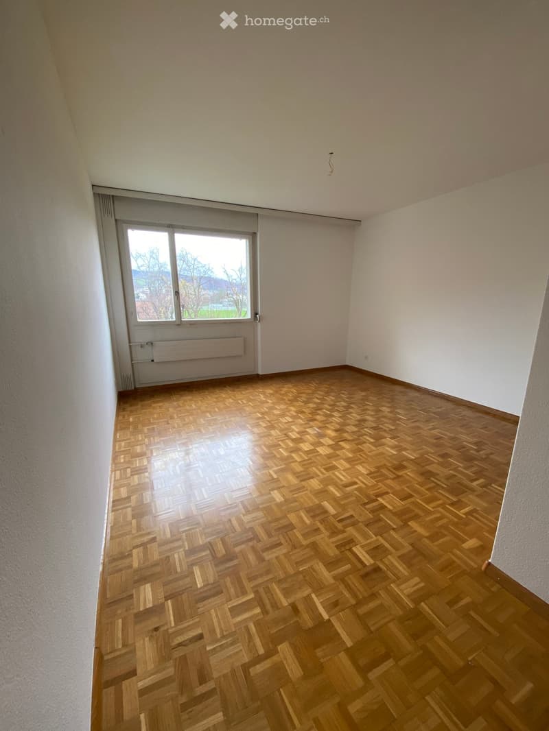 Empty room with wooden herringbone flooring, white walls, and a large window with a view of the outdoors.