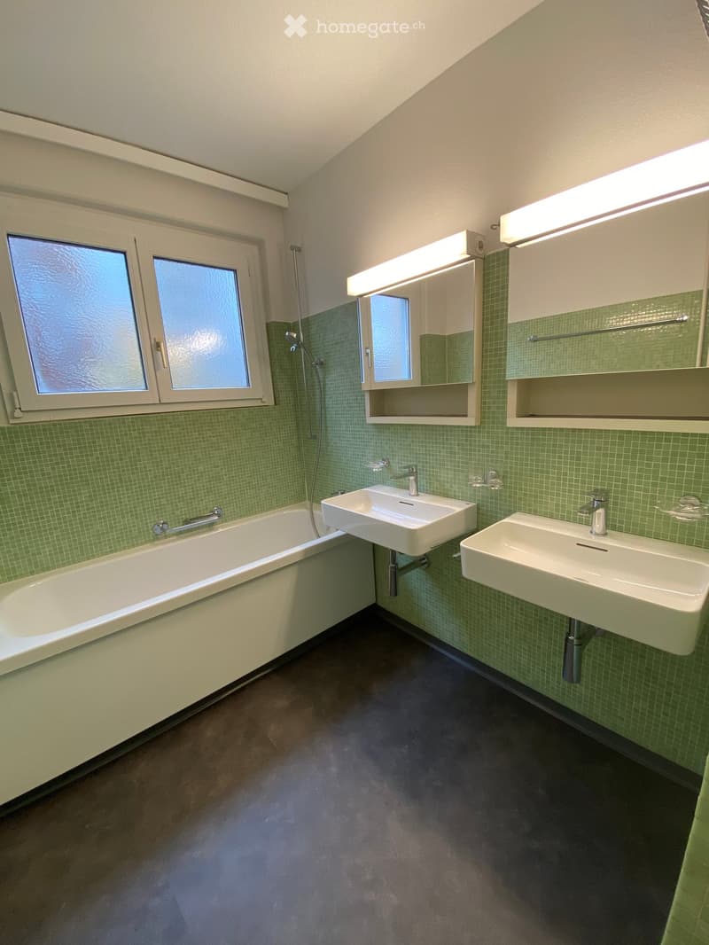 Modern bathroom with green tiled walls, a bathtub, two sinks, a mirror, and frosted windows.