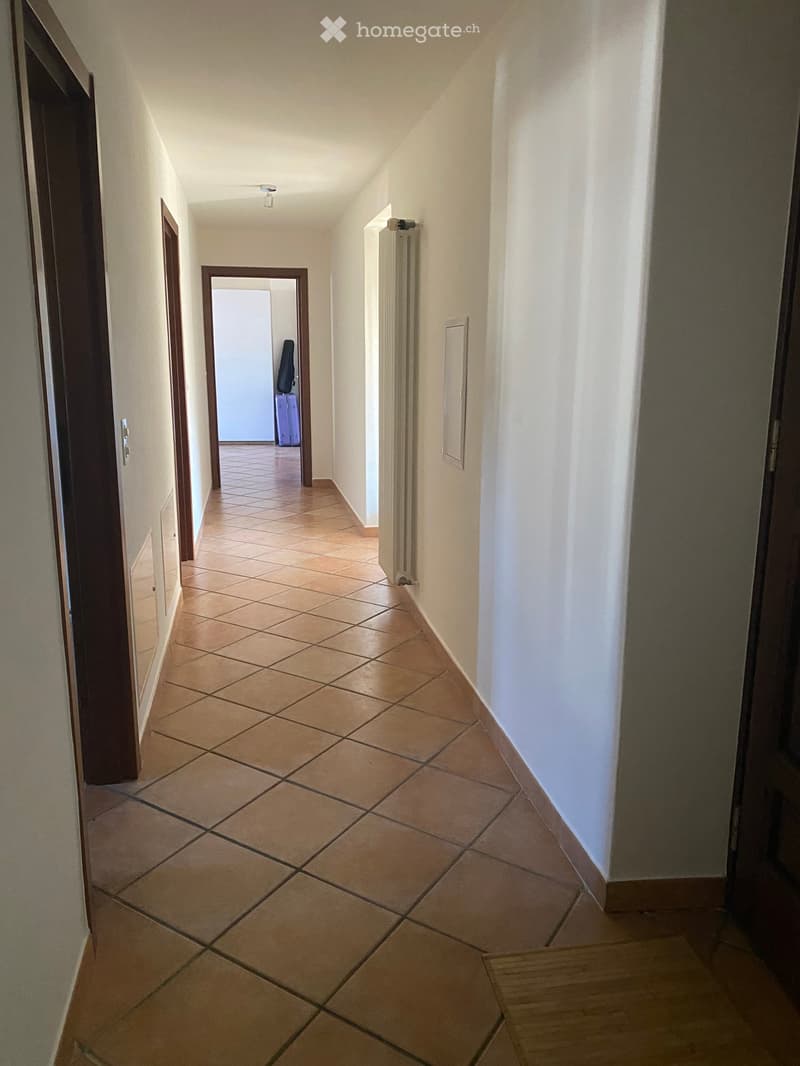 A narrow hallway with tiled flooring, white walls, and several doors leading to other rooms. A radiator is visible on one wall, and a small piece of furniture is seen in an adjacent room.