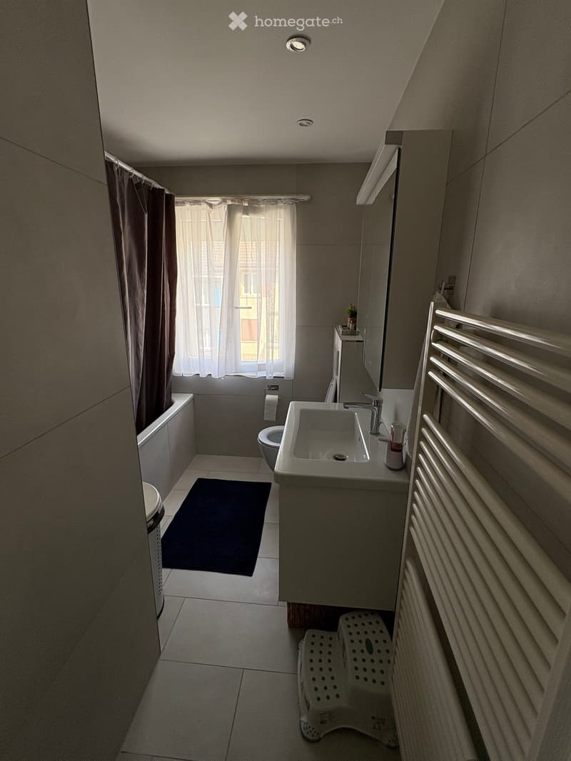 Modern bathroom with a sink, toilet, bathtub, radiator, and a small stool near the sink. The room features a window with sheer curtains and a dark bathmat on the floor.
