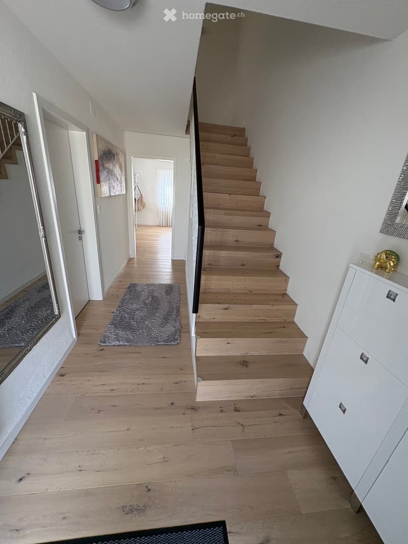 A modern hallway with wooden flooring, a staircase leading upwards, and a small grey rug placed near the stairs. The walls are painted white, and there are mirrors and a small cabinet visible on the right side.
