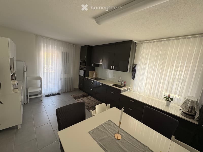 Modern kitchen with dark cabinets, white countertops, a dining area with a table and chairs, and a small bar area with a stool. The space features large windows with sheer curtains and a tiled floor.