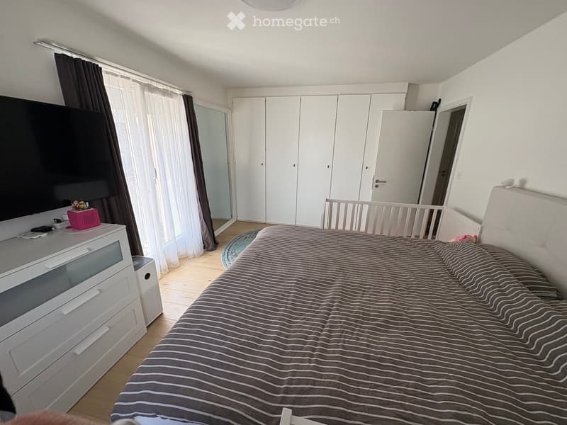 Modern bedroom with a large bed, striped bedding, white wardrobe, TV on a stand, and large windows with curtains.
