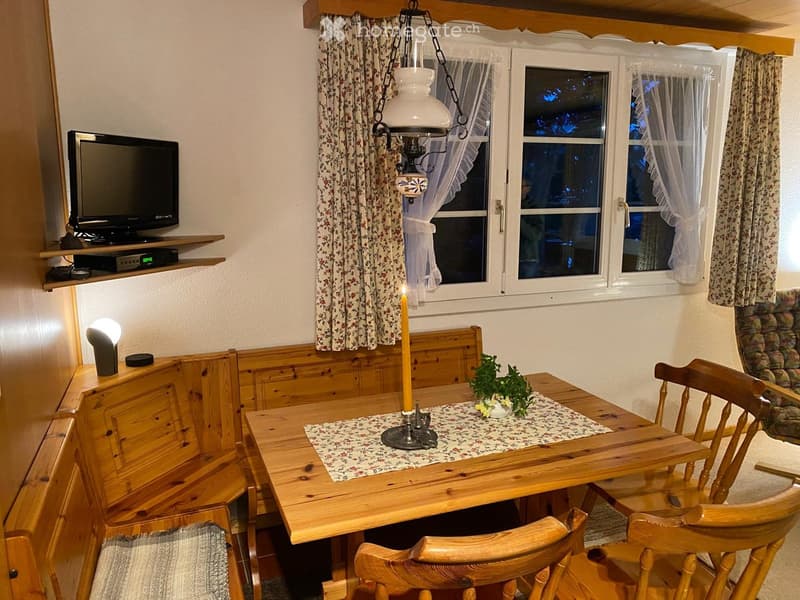 Cozy dining area with wooden table, chairs, bench seating, television on shelf, candle, and flower pot.