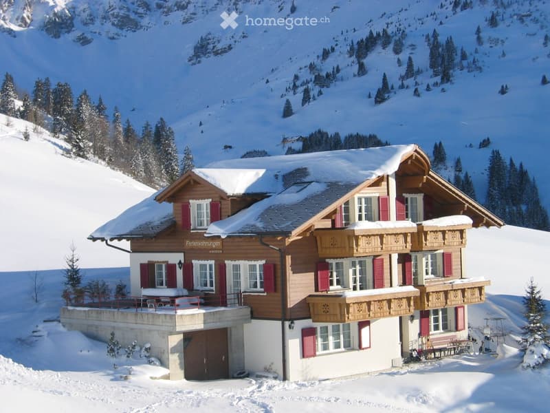 A large wooden house with red shutters and white trim, situated in a snowy mountain landscape. The house features multiple windows, balconies, and a garage entrance.