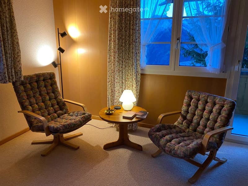 Cozy living room with two patterned armchairs, a small round wooden table with a lamp, books, and a candle, illuminated by warm lighting and large windows with floral curtains.