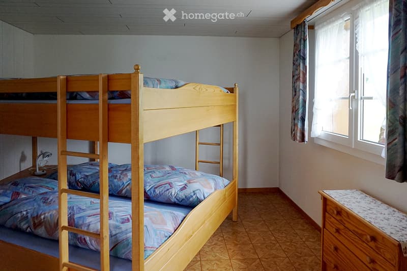Cozy bedroom with a wooden bunk bed, colorful bedding, a small nightstand with a lamp, and a window with curtains.