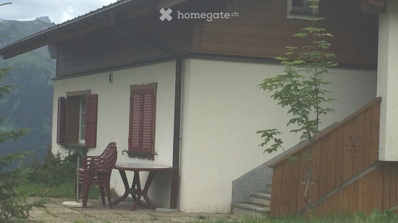 Two-story house with white walls, red shutters, and a wooden staircase leading to the entrance. A small table and chairs are placed near the windows.