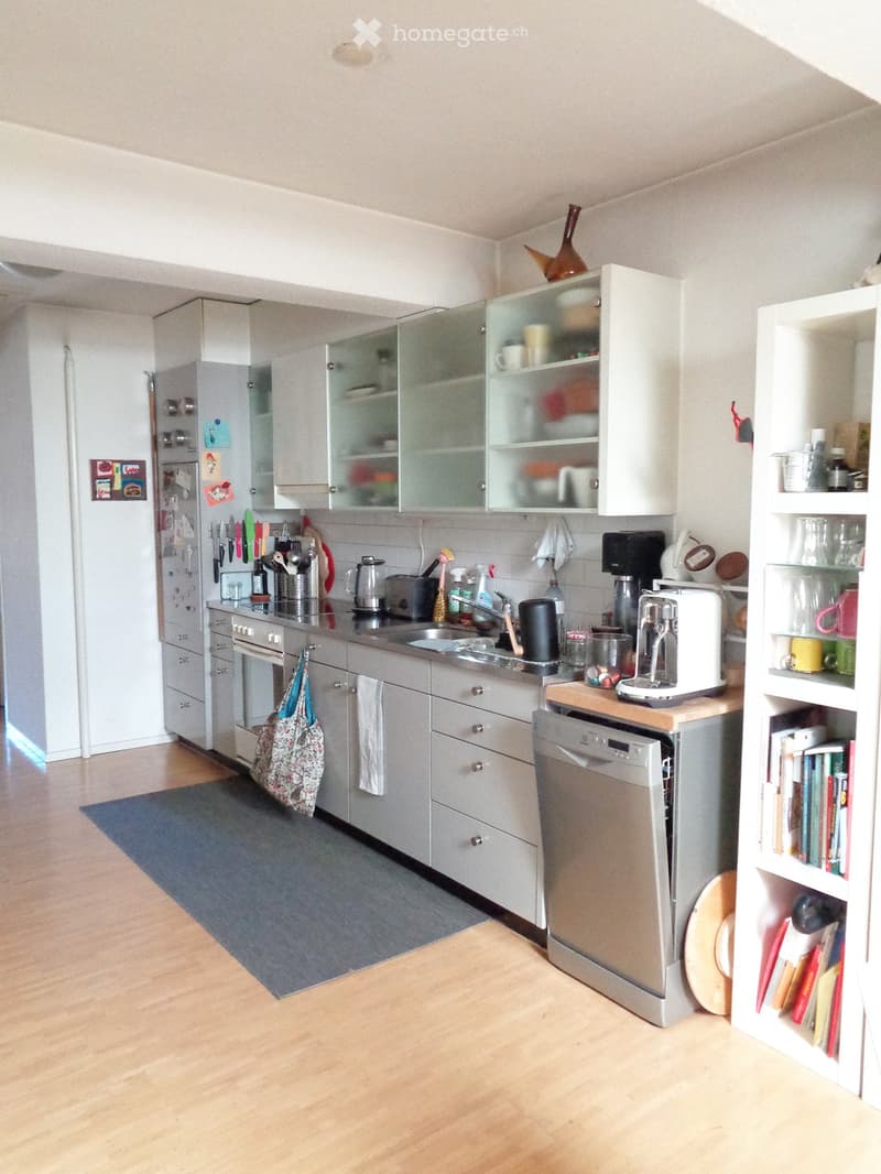 Modern kitchen with grey cabinets, stainless steel appliances, wooden floor, and white shelving unit with books and decorative items.