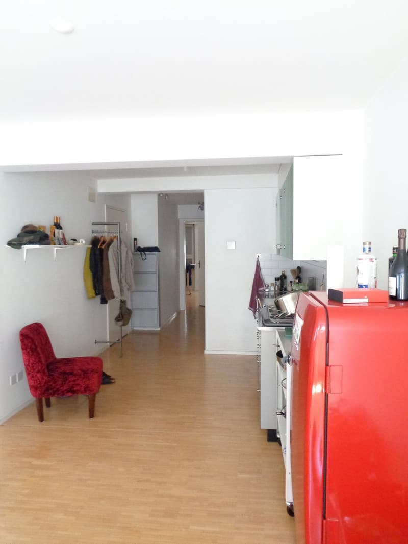 A modern, open-plan living and kitchen area with light wooden flooring, white walls, a red chair, a red refrigerator, and various bottles and items on the counter and shelves.