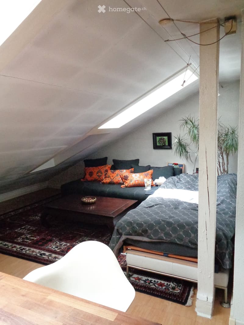 Cozy attic room with a bed, sofa, coffee table, and a white chair. The room features wooden flooring, a skylight, and decorative rugs.