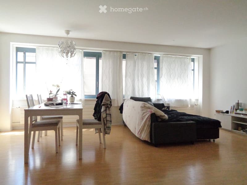 A well-lit room with a sofa, a dining table with chairs, a chandelier, and large windows with curtains. The floor is wooden, and there are decorative items on the table and shelves.