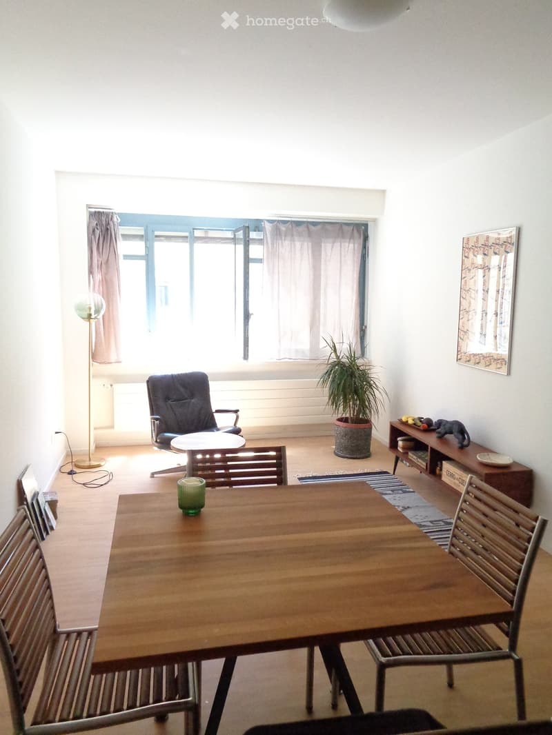 Bright living room with wooden floor, large window, sofa, dining table, chairs, plant, and artwork on wall.