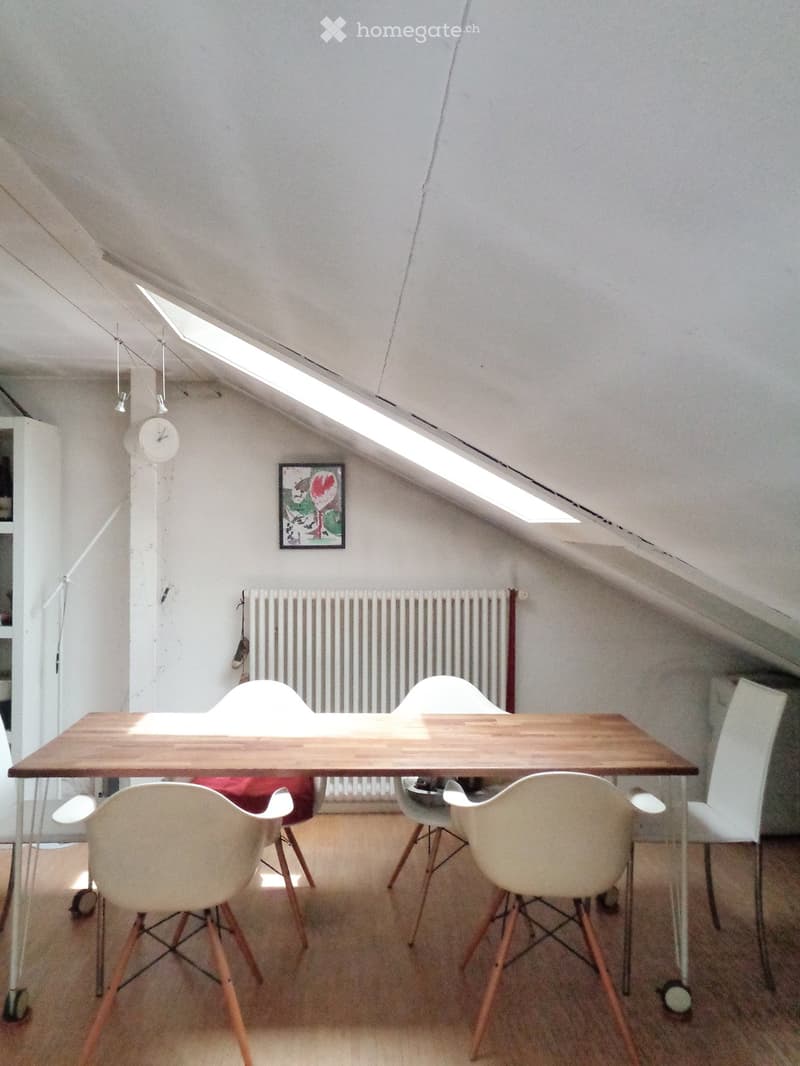 Bright attic room with wooden table, white chairs, radiator, clock, and picture frame on the wall.
