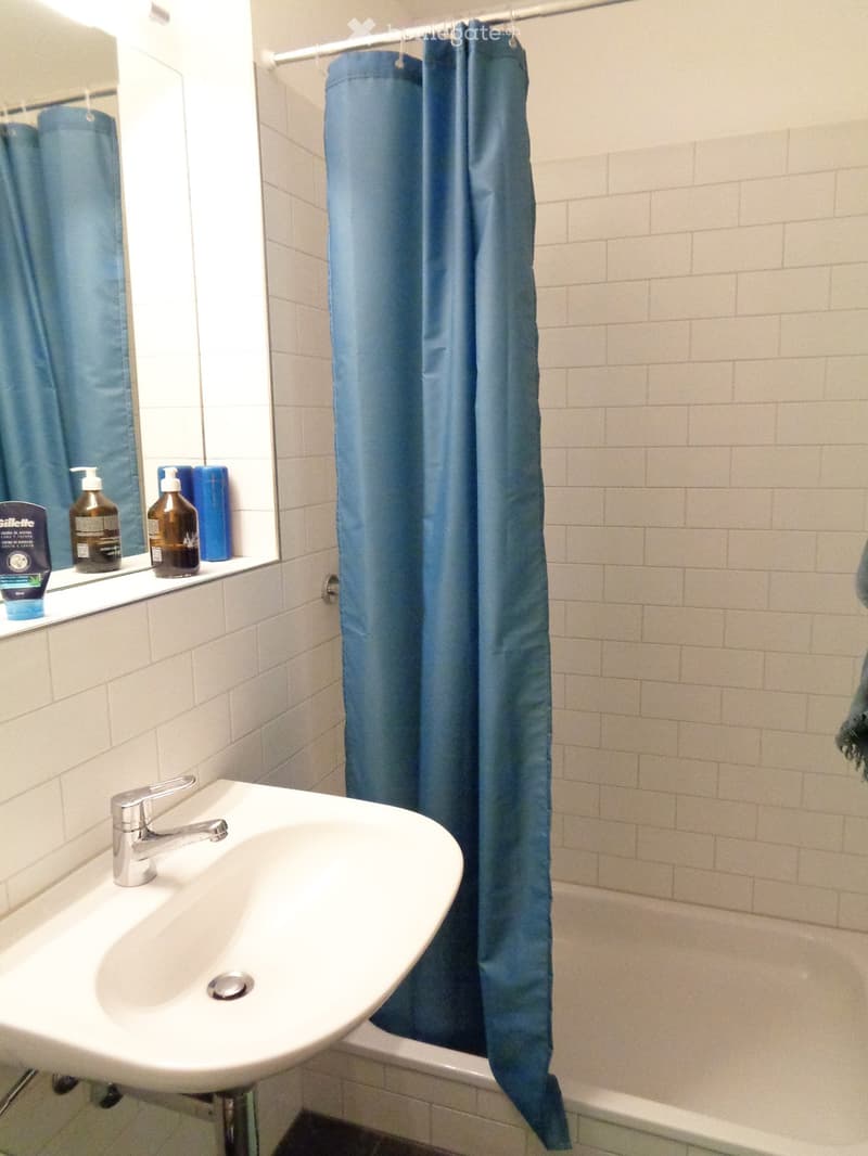 Bathroom with a white sink, chrome faucet, mirror, and blue shower curtain. Several bottles of toiletries are placed on the windowsill.
