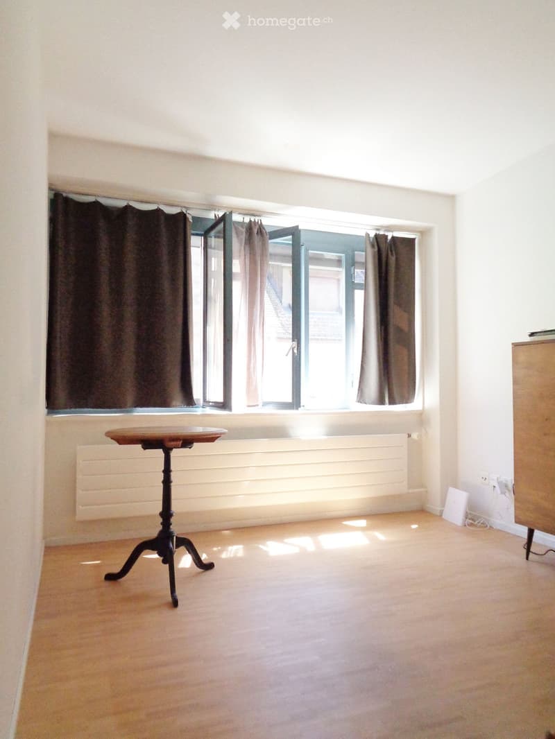 Empty room with wooden floor, large window with curtains, radiator, and a small table near the window.