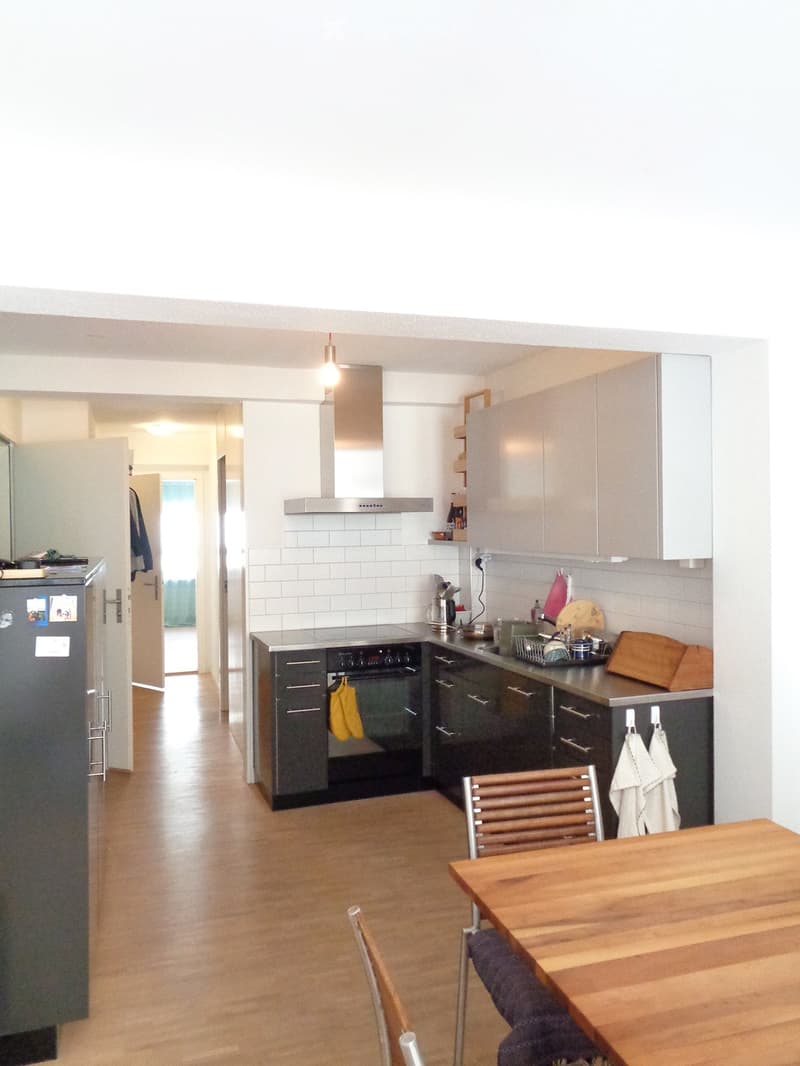Modern kitchen with light-colored cabinets, stainless steel appliances, wooden flooring, and a small dining area with a table and chairs.