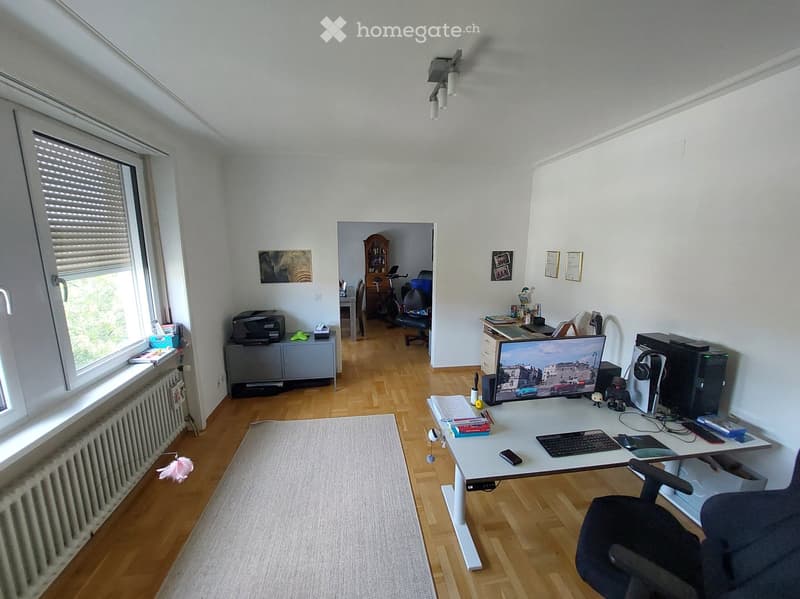 A well-lit room with wooden flooring, a large desk with a computer setup, a printer, and a comfortable chair. There is a window with blinds, a radiator, and a small rug on the floor. The room also features a doorway leading to another area with a dining t