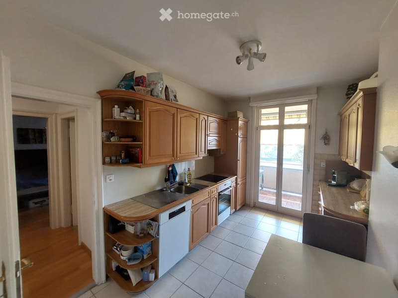 Well-lit kitchen with wooden cabinets, tiled floor, and a large window leading to a balcony. Includes a sink, oven, dishwasher, shelves, and a countertop.