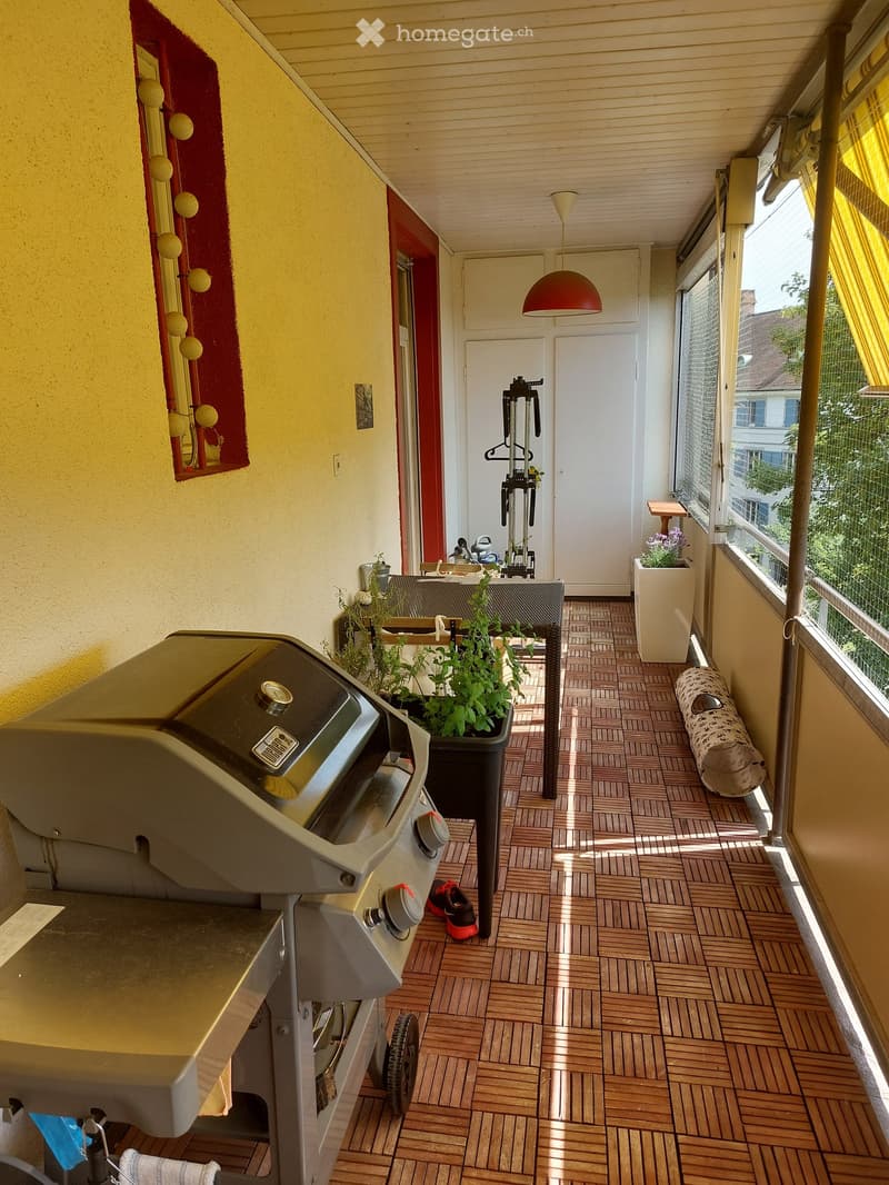 Balcony with wooden flooring, grill, small table, chairs, potted plants, and a pet bed. Features large windows with yellow curtains and a hanging lamp.
