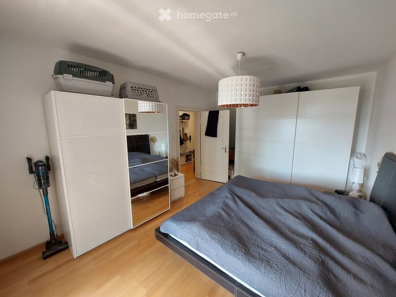 Well-lit bedroom with a large bed, modern wardrobe, and a hanging lamp. The room features wooden flooring and a visible adjacent space through an open door.