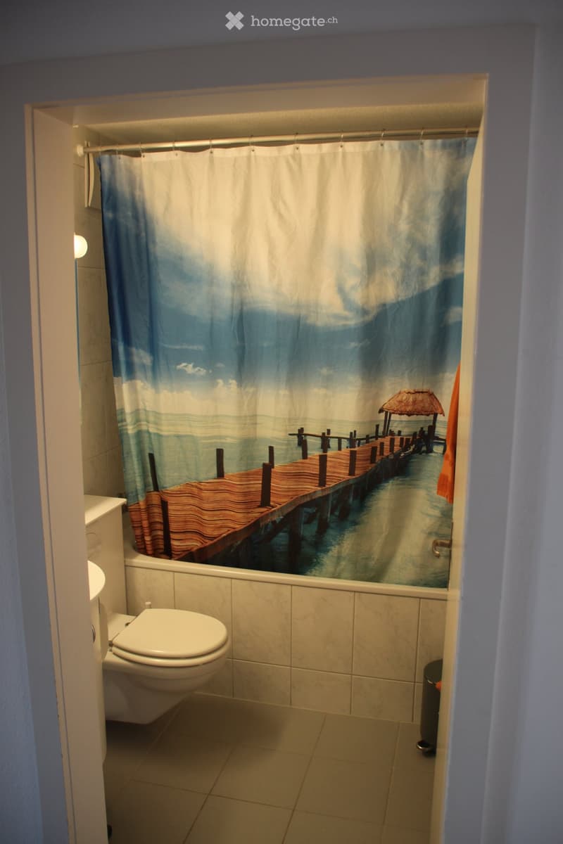 Bathroom with a toilet, bathtub, and a decorative shower curtain featuring a beach scene with a pier and thatched-roof structure. Light-colored tiles on walls and floor.