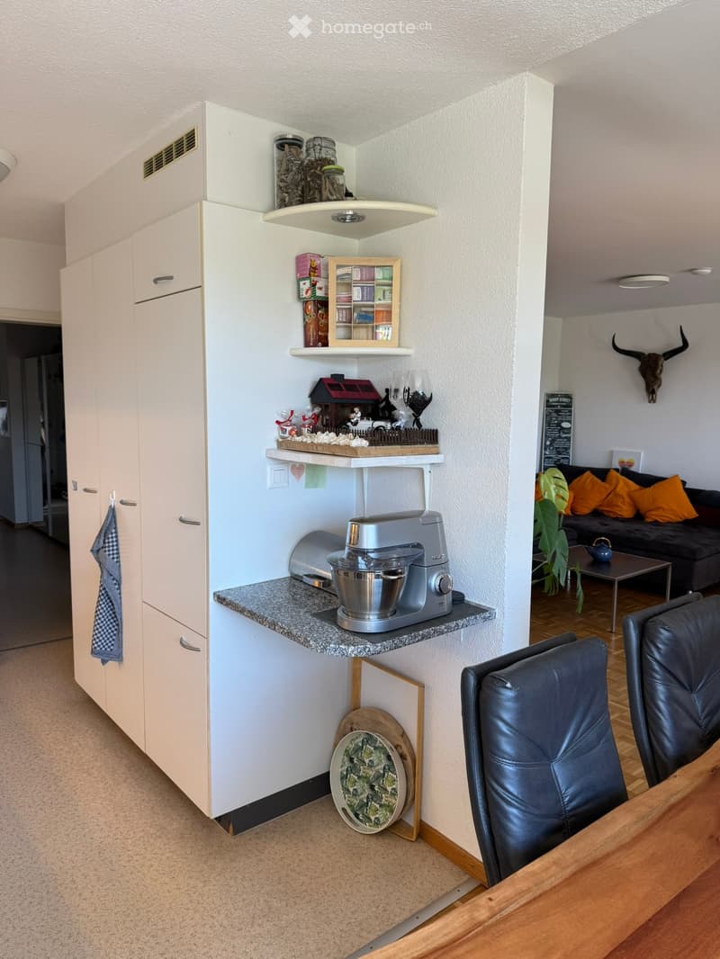 A modern, open-plan living and kitchen area with white cabinets, a granite countertop, a mixer, shelves with decorative items, a sofa with orange cushions, a small table, and a skull on the wall.