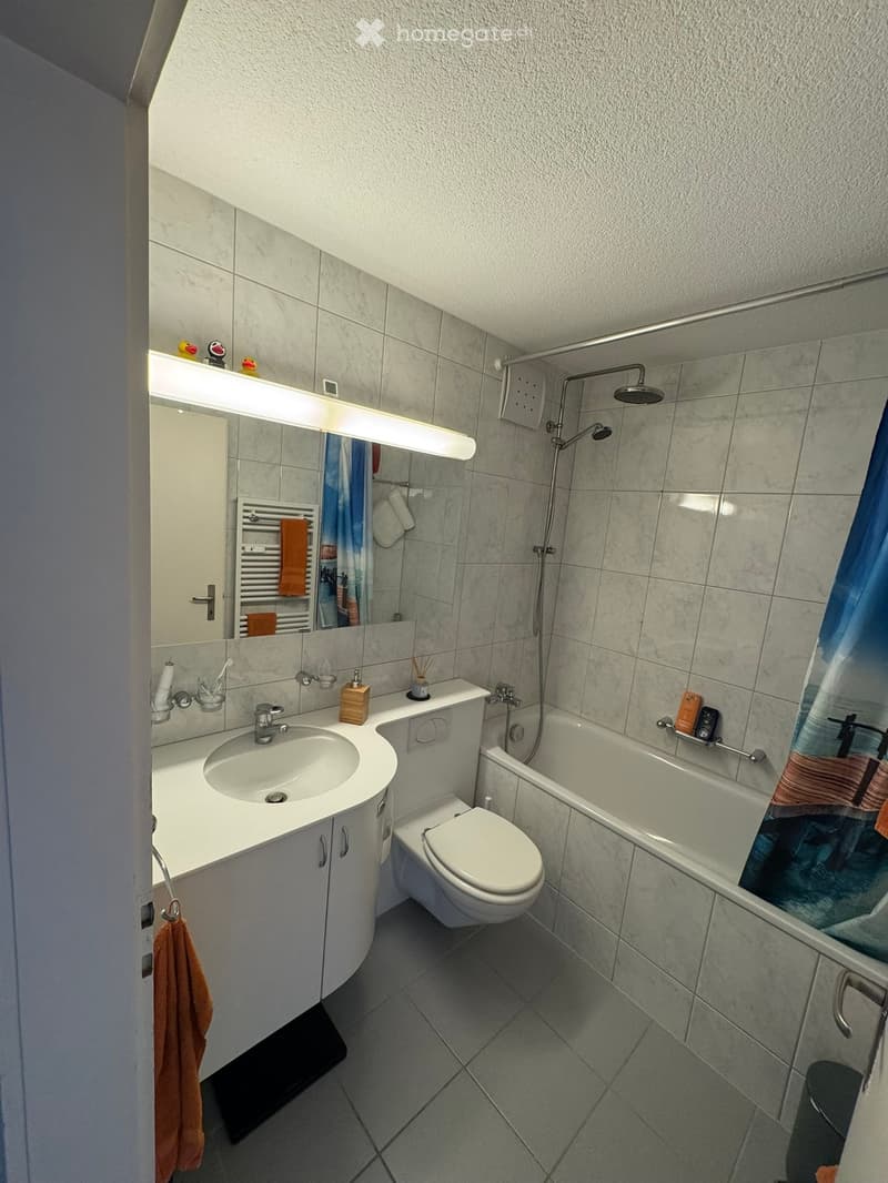 Modern bathroom with a white sink, toilet, bathtub, and large mirror. Features tiled walls and floor, a towel rack, and a showerhead.