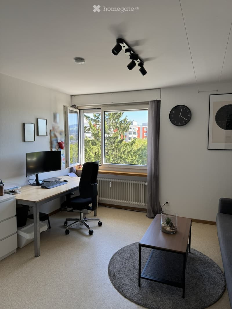 A modern, well-lit room with a desk setup, a comfortable chair, a small table with a plant, and a sofa. Large windows provide natural light and a view of greenery outside.