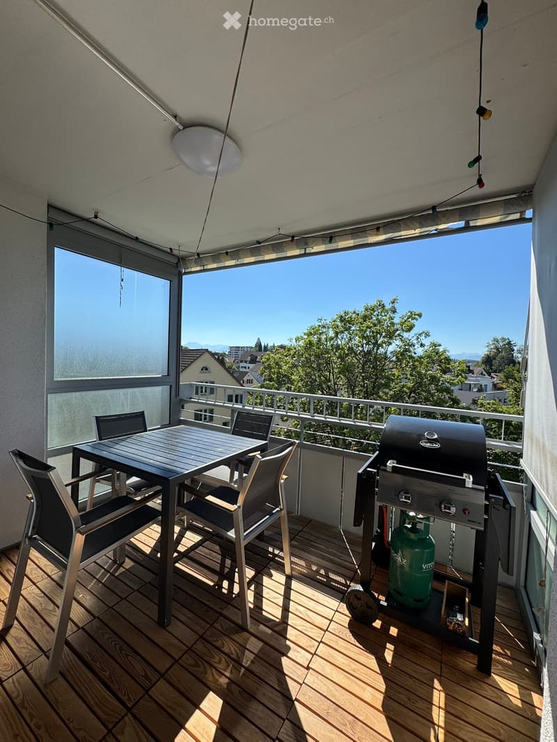 Balcony with wooden decking, table and chairs, grill, and scenic view of trees and houses.