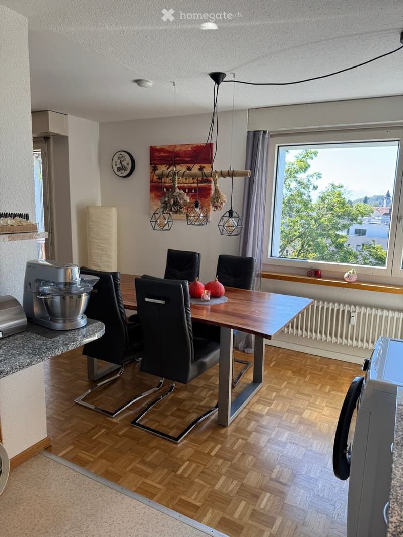 Modern dining area with wooden herringbone flooring, a large wooden dining table surrounded by black leather chairs, a mixer on the counter, and a large window with a view of greenery.